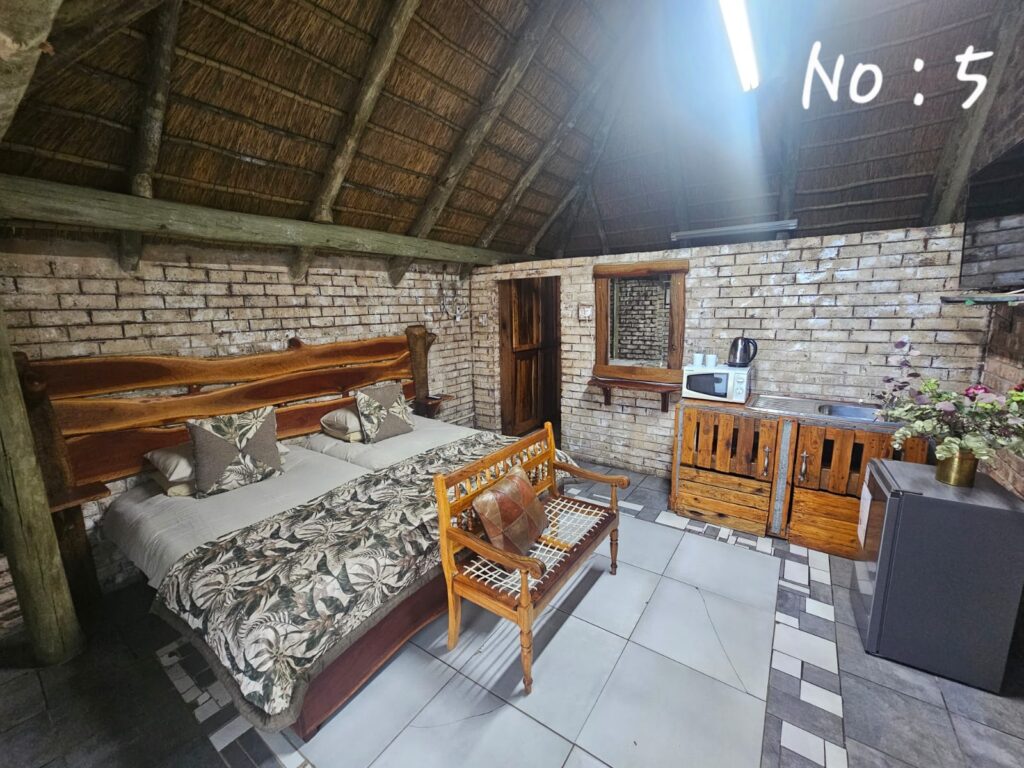 Self-catering room with brick walls, thatched ceiling, bed, kitchenette, and wooden bench.
