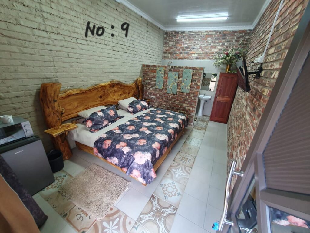 Large bed with floral linens, a massive raw-wood headboard, patterned floor tiles, and a brick partition wall leading to the bathroom.